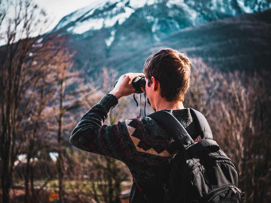 A young man with a backpack uses binoculars to admire a scenic mountain view.